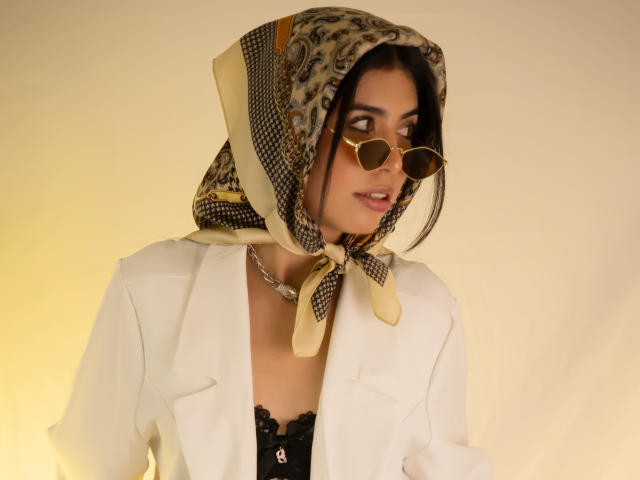 MiluSky with a patterned headscarf, gold heart-shaped sunglasses, white blazer, black lace top, and chain necklace, looking to the side, with a subtle, sexy smile.
