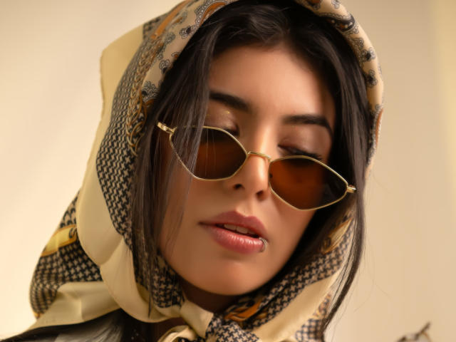 MiluSky with brown aviator sunglasses, tan patterned headscarf, and closed eyes, has smooth, tan skin, and full pink lips. Her long black hair is slightly visible.