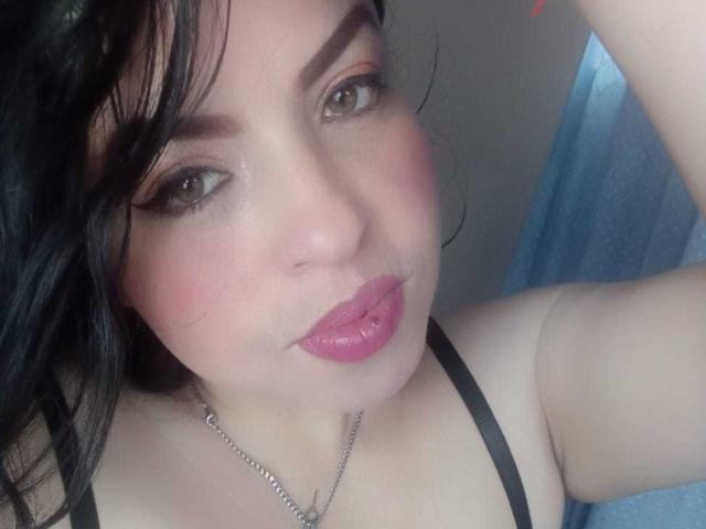 Close-up selfie of PaulaEllie with fair skin and black hair, slightly wavy. She has dark, well-defined eyebrows, light brown eyes, and pink blush on her cheeks. Her lips are full and painted a bright pink. She wears a thin black strap, possibly a bra, and a silver necklace. Her right arm is raised, and the blue curtain in the background is blurred. The image has a slightly glossy texture, emphasizing her skin's smoothness. Her expression is neutral, with a slight pout on her lips.