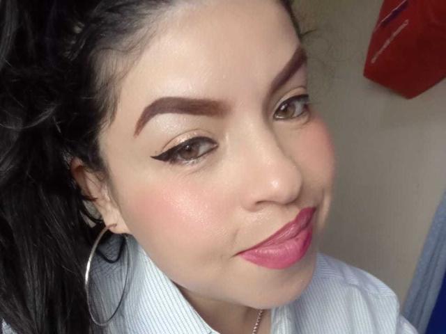 Close-up of PaulaEllie with medium brown skin, dark eyebrows, and thick eyeliner. She has pink lipstick, a slight smile, and wears a light blue striped shirt with large silver hoop earrings. Her black hair is tied back, and she has a subtle glow on her cheeks. The image focuses on her face, with a red object in the upper right corner.
