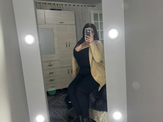 VenusxMusa met grote borsten, gekleed in een zwarte jurk, zwarte leggings en een beige vest, zittend voor een spiegel, met een telefoon in de hand, met heldere lichten die reflecteren, in een slaapkamer..