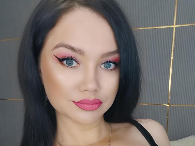 KimHonney has blue eyes, black hair, pink eyeliner, red eyeshadow, and pink lipstick. She's wearing a black top and has a cute, sexy face with a subtle smile.