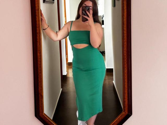 VicluX in a green, cut-out, form-fitting dress, showing her tits and ass, taking a mirror selfie with one hand on the wall, wearing white socks.