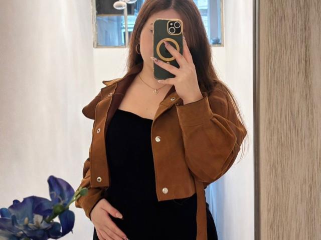 VicluX com cabelo castanho, vestido preto e jaqueta marrom, segurando o celular, mostrando o bumbum, em pé, selfie no espelho, com uma flor azul no lado esquerdo..