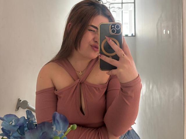 VicluX with brown hair, wearing a pink off-shoulder top, taking a mirror selfie with a gold ring necklace, holding a phone with a ring case. Boobs visible.