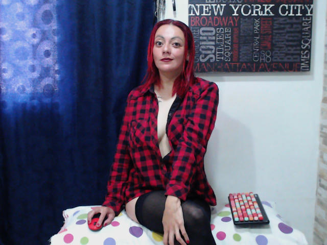 SamanthaSwan with red hair, wearing an open red and black checkered shirt, black thigh-highs, sitting on a polka-dotted bed, looking up, with a remote control beside her.
