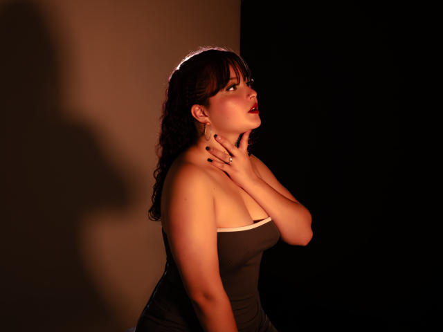 GiaDonovan, con cabello castaño rizado, lápiz labial rojo y vestido negro, tocándose el cuello, sombra en la pared, mirando hacia arriba, iluminación suave, sus senos ligeramente visibles, fondo oscuro..