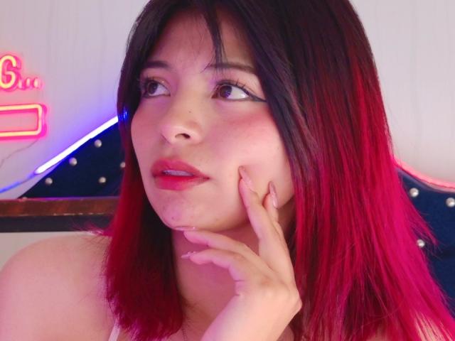 ShofiaWett with red-to-black ombre hair, pink eyeliner, and red lipstick, touching her cheek with her right hand, looking to the side, pink neon light in the background.