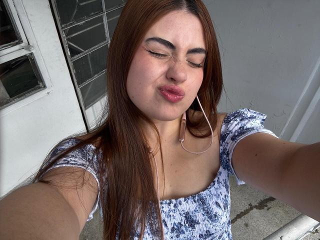 VicluX a les lèvres roses et gonflées, les yeux fermés et les cheveux bruns. Elle porte un haut bleu et blanc à motifs et a des écouteurs. Ses seins sont petits et elle prend un selfie..