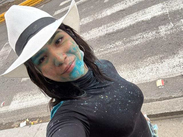 ScarlletZhen, con pintura azul en la cara, un sombrero blanco, una blusa negra y los senos visibles, está de pie en una calle, mirando a la cámara, con aros grandes y sonriendo ligeramente..