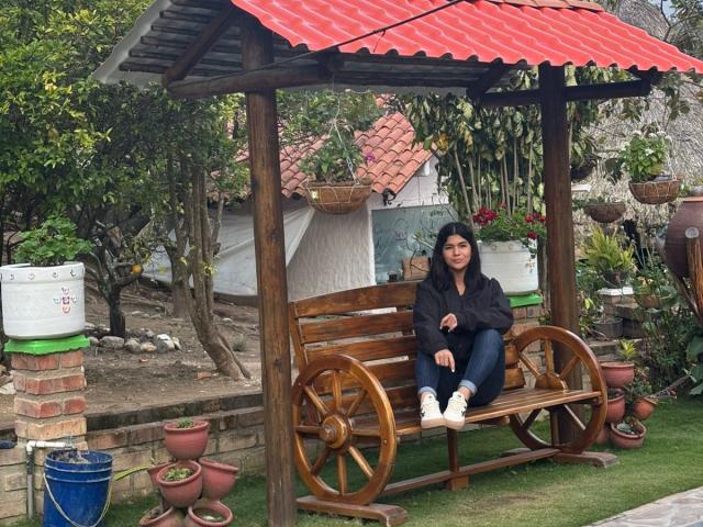 ScarlletZhen sits on a wooden bench with wheels, wearing a black jacket and blue jeans, under a red metal roof, surrounded by potted plants and green grass.