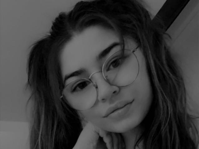AnnaGorgeous has wavy, messy hair, wearing round glasses, and is looking at the camera with a slight smile, resting her chin on her hand. Black-and-white selfie.