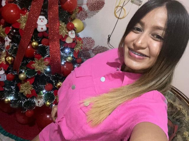 AlondraLira with long brown hair, pink shirt, smiling, standing next to a Christmas tree with red ornaments and a snowman decoration, showing her cute face and small tits.