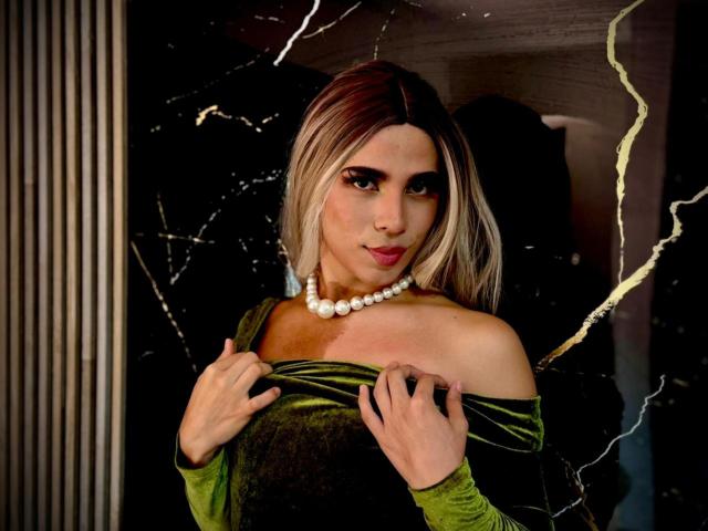 LyaCollins with blonde hair, green off-shoulder dress, holding it down, pearl necklace, dark background, looking at camera, slightly parted lips, subtle makeup, fingers near collarbone.