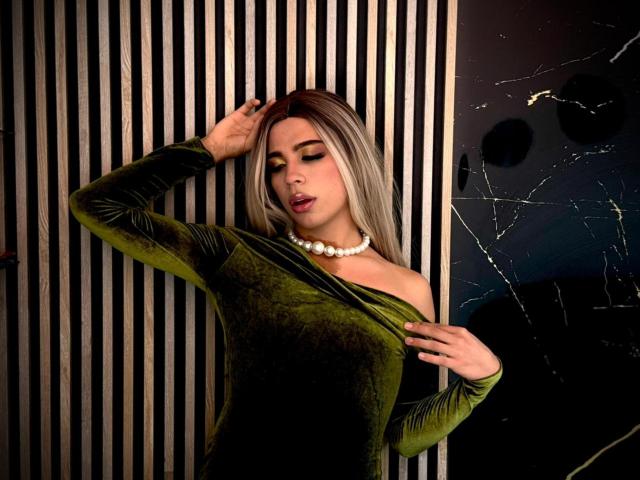LyaCollins with blonde hair, green velvet dress, pearl necklace, golden eyeshadow, hand in hair, ass slightly visible, sexy, confident, right hand on dress, left hand on head.