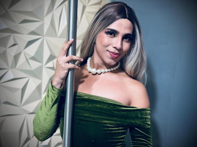 LyaCollins with blonde hair, green off-shoulder dress, white pearl necklace, holding pole, smiling, showing her tits, fair skin, medium build, standing, posing, sexy, confident.