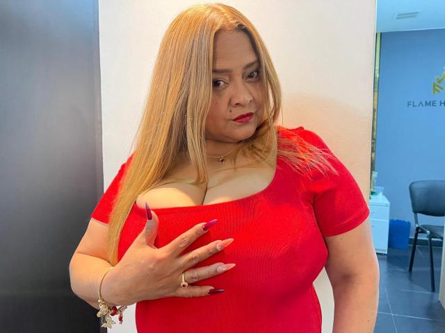 MonzerratBoyer with blonde hair, red dress, and red lipstick, touching her big boobs with purple nails, wearing a necklace and ring, looking down with a slight smile.