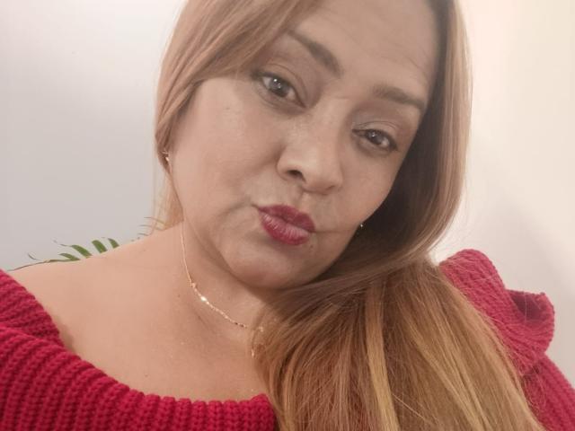 MonzerratBoyer with pouty red lips, wearing a red knitted top, has light brown hair, a gold necklace, and a slight smile, showing her cute ass face.