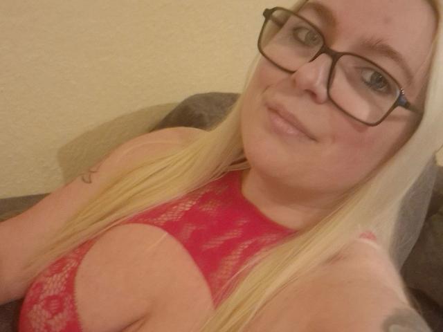 A selfie of Sweet-Butterfly-hot with blonde hair, glasses, and a pink lace bra showing big tits, smiling slightly, and sitting on a grey couch.