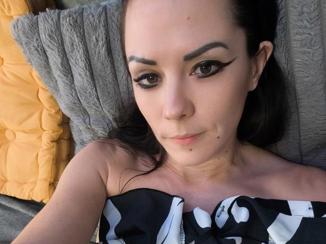 OrianaLaFrancaise lies on a textured gray pillow, head resting on a yellow pillow. She has dark brown hair, thick, well-groomed eyebrows, and dramatic black eyeliner. Her skin is light with a small mole under her right cheek. She wears a strapless black and white patterned top, revealing her bare shoulders. The photograph is close-up, focusing on her face and upper chest. Her expression is neutral, with a slight, almost imperceptible smile. The image has a realistic, intimate style.