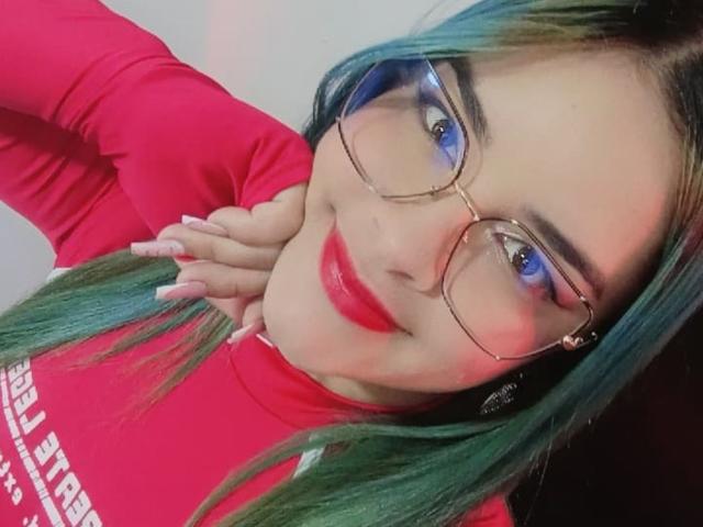 AuroraSantorini met groen-blauw haar, rode lipstick en een bril, lachend naar de camera, gekleed in een rode blouse met lange mouwen, met haar hand op haar nek, en haar schattige achterkant showend..