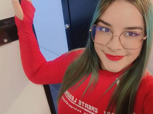 AuroraSantorini met groen haar, rode lippenstift en een bril, draagt een rode blouse met lange mouwen, glimlacht en heft haar rechterarm, waarbij haar borsten lichtelijk zichtbaar zijn..