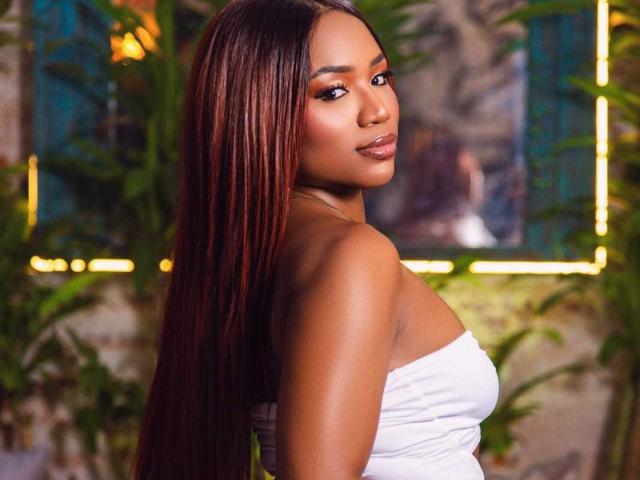 FarinaLyte has long, straight, dark auburn hair flowing past her shoulders. She wears a strapless white top, exposing her shoulders and upper back. Her skin is smooth and dark brown, with subtle makeup highlighting her eyes and lips. She looks over her shoulder with a calm, confident expression. The background is blurred with green plants and yellow neon lights, creating a contrast with her white top and dark hair. The image uses warm, natural lighting.