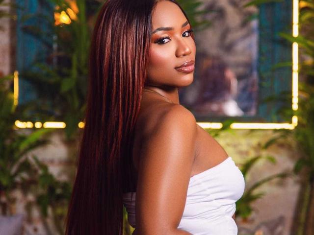 FarinaLyte stands with her back slightly turned, looking over her shoulder. She has long, straight, dark auburn hair that cascades down her back. She wears a white strapless top that highlights her smooth, dark brown skin. Her makeup is bold, with shimmering eyeshadow and glossy pink lips. The background is blurred with greenery and yellow lights. Her confident, relaxed expression adds to her overall striking appearance.