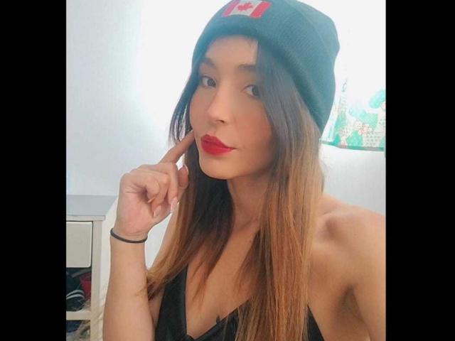 MindyLove has long brown hair, red lipstick, and a black beanie with a red logo, touching her chin with a finger, wearing a black tank top, showing her tits.