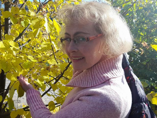 SunnySoul-hot with short blonde hair, pink glasses, and pink turtleneck, smiling at yellow leaves, holding a black backpack with red patterns. She's looking over her shoulder.