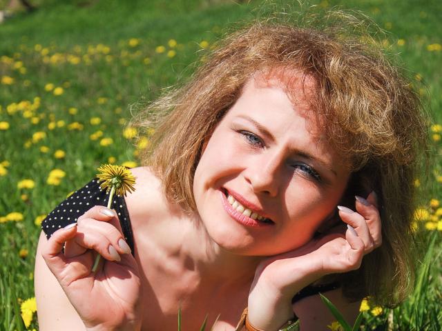 SunnySoul-hot with curly blonde hair, holding a dandelion, smiling, lying in green grass with yellow flowers, wearing a black polka dot top, looking up with blue eyes.