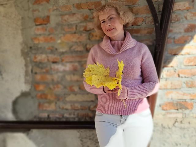 SunnySoul-hot with curly blonde hair, pink turtleneck sweater, white pants, holding yellow leaves, standing against brick wall, smiling softly, hands gently holding leaves, soft natural light.
