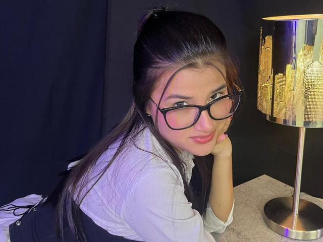 Iris Blair, con lunghi capelli neri, occhiali, camicia bianca, gilet nero, appoggiata al tavolo, con la testa appoggiata sulla mano, accanto a un lampione che illumina una veduta cittadina, guarda la fotocamera con un leggero sorriso..