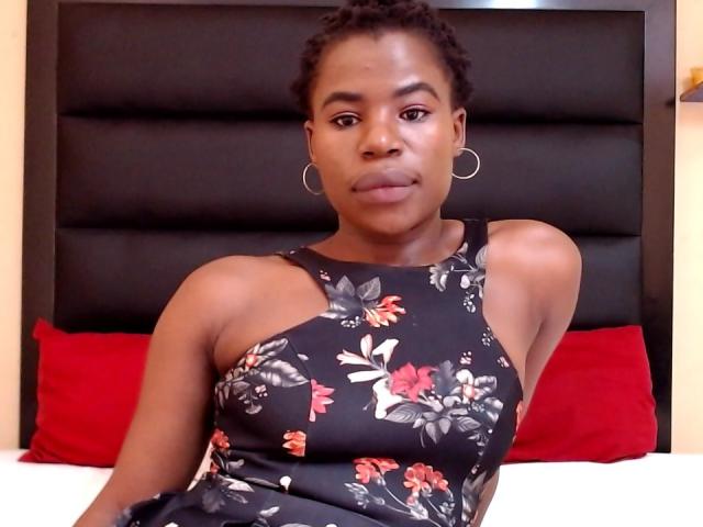 RoundyAss with short curly hair, wearing a black floral dress, silver hoop earrings, sitting on a bed with red pillows, has a serious expression, medium-sized tits, dark brown skin.