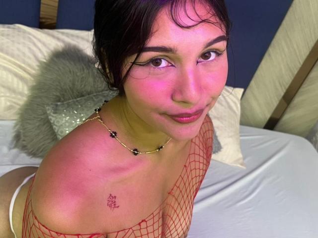 ValeFox, avec ses joues roses, ses cheveux foncés et son top en résille, regarde vers le haut, montrant de petites seins et un tatouage sur son épaule, portant un collier de perles noires, sur un lit..
