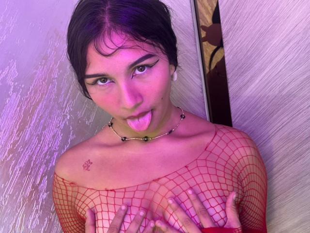 ValeFox, avec ses cheveux foncés, son maquillage violet et sa langue tirée, portant un haut en filet rouge, cachant ses petites seins avec ses mains, et un collier de perles noires..