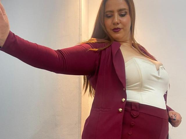 Freya, mit langen braunen Haaren, trägt einen dunkelroten Blazer, ein weißes Top ohne Träger und eine dunkelrote Hose mit hoher Taille. Ihr rechter Arm ist ausgestreckt, wodurch ihre Brüste sichtbar werden..