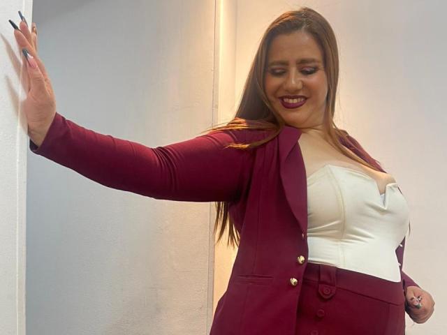 FreyaSeid with long brown hair, red blazer, white strapless top, and red skirt, smiling, hand on wall, showing off her tits and small ass, dark nail polish.