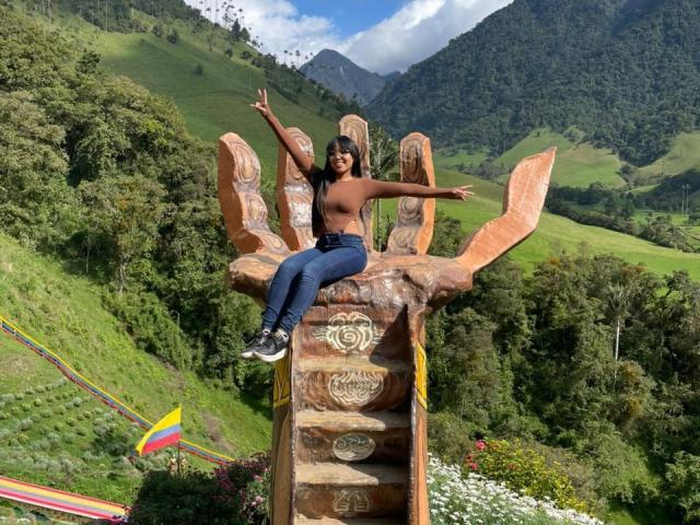Heyly, topless con la pelle scura, jeans blu e scarpe da ginnastica bianche, si siede su una gigantesca statua di legno con le braccia aperte, sfondo di montagna.