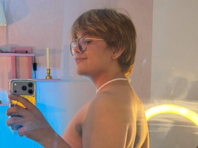 BenjiVampire with short brown hair, glasses, and a pearl necklace, taking a selfie with a yellow phone, showing bare shoulders and a subtle smile. Boobs not visible.