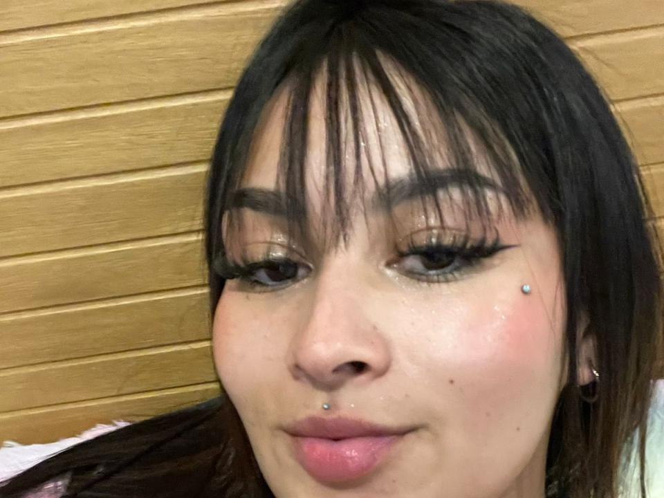 GigiFoles has black hair with bangs, heavy eyeliner, and a nose piercing with a small blue stud. She's wearing pink lipstick and has a lip piercing.