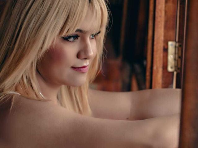 Evees with blonde hair and bangs, looking sideways with a slight smile, showing her tits and bare shoulders, standing by a wooden door. Soft light on her face.