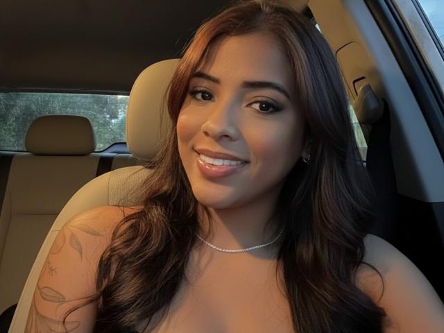 JuMarques, brown-haired, smiling with glossy pink lips, wearing a necklace, has tattoos on her right arm, sitting in a car, showing off her cute, curvy tits.