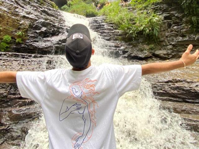Georginasim with arms out, facing rocky waterfall, wearing black cap, white t-shirt with line art of topless woman, standing in front of cascading water.