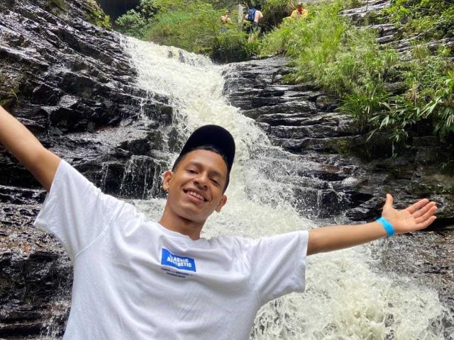 Georginasim, smiling with arms out, stands in front of a waterfall. Wearing a white shirt and black cap, has a blue wristband. Water cascades over dark rocks.