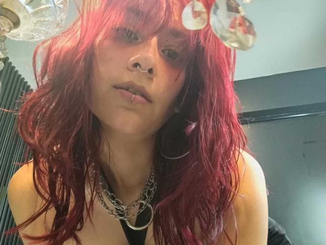 LolyYunno with red, wavy hair, nose ring, lip piercing, and chain necklace, wearing a black tank top, looking down at the camera. Her tits are partially visible.