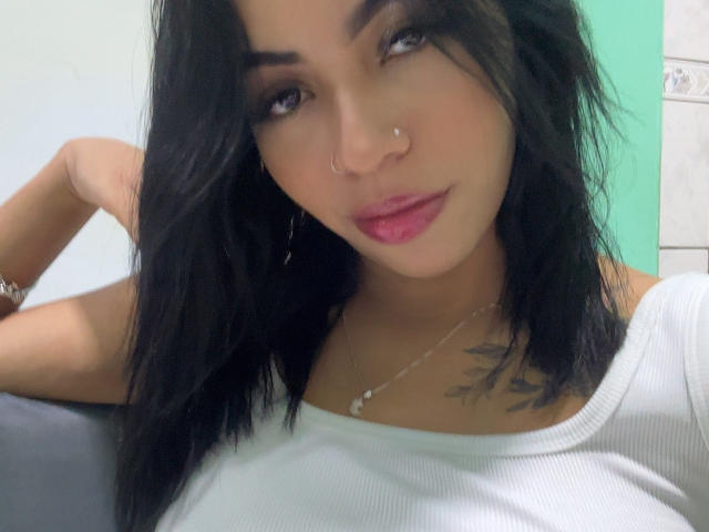 FelinaRara with black hair, nose ring, and pink lipstick, wearing a white ribbed top, showing a tattoo on her chest, and a necklace with a small pendant.