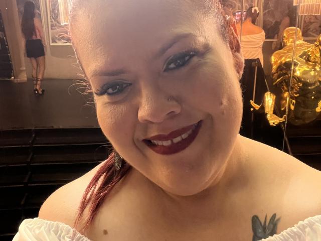 VanesaMoka with red hair, white off-shoulder top, dark lipstick, nose ring, smiling, visible butterfly tattoo on right chest, gold statue in background.