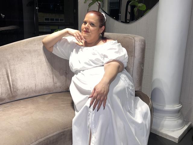 VanesaMoka is sitting on a beige sofa, wearing a white dress, with red nails, and a relaxed expression, her right hand touching her chin, her left on her thigh.