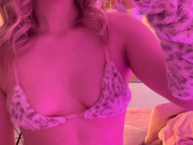 A close-up of Synrix with pink lighting. Synrix has wavy blonde hair, wearing a furry, leopard-patterned bralette that slightly reveals their breasts. The right arm is raised, holding a fuzzy garment. Synrix's skin is light, and the image has a warm, pinkish hue. The bralette is textured with small, dark spots on a white background. The focus is on the upper body, with no visible background details. The image is candid and has a casual, intimate feel.